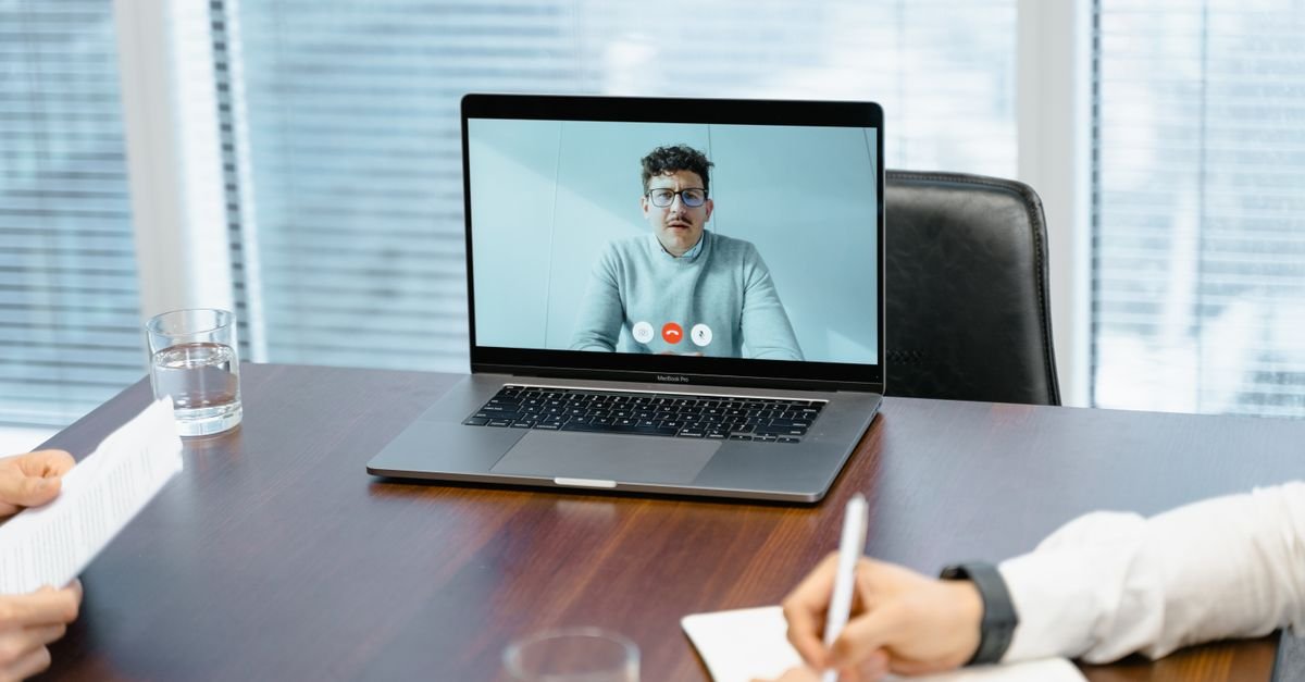 Notebook sobre mesa de reunião exibe participante em chamada de vídeo enquanto duas pessoas tomam notas